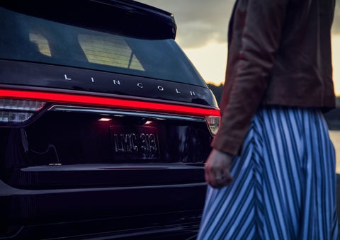 A person is shown near the rear of a 2024 Lincoln Aviator® SUV as the Lincoln Embrace illuminates the rear lights | Doggett Lincoln of Beaumont in Beaumont TX